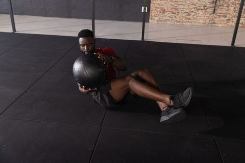 African american man performing seated medicine ball russian twist on rubber gym floor