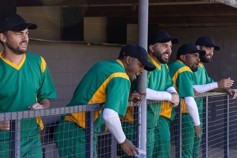 Baseball teammates leaning on dugout fence laughing and cheering in green uniforms during game