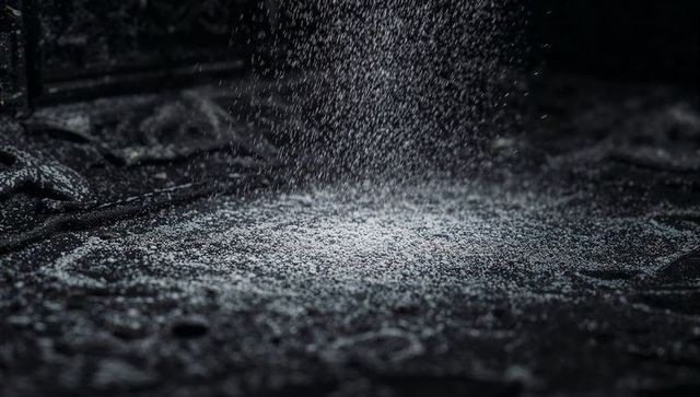 Fine white granules sprinkling on dark fabric, dramatic overhead lighting, macro texture