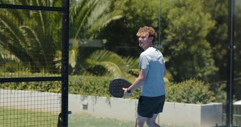 Male player looking back holding paddle on sunlit outdoor pickleball court with glass wall
