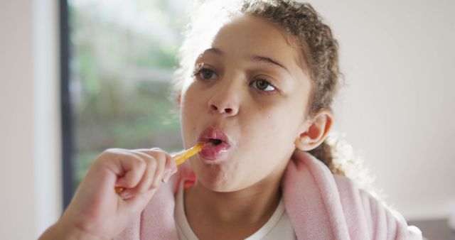 Young girl with afro hair brushing her teeth in soft morning light. She is draped in a pink robe, emphasizing the early morning routine. The scene portrays the importance of dental hygiene in everyday life. Ideal for use in health and dental care promotions, educational content about children’s health routines, and family lifestyle advertising.