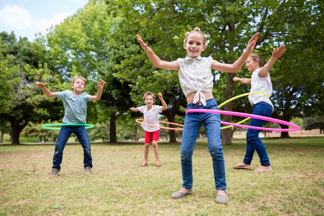 Children enjoying a sunny day in the park while playing with hula hoops. Ideal for use in advertisements promoting outdoor activities, healthy lifestyles, and children's fitness. Can also be used in educational materials about physical activity and group play.