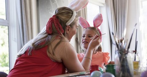 Mother painting daughter's face wearing bunny ears during spring crafting and playtime at home