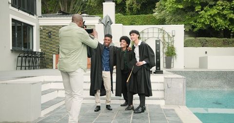 Proud father photographing sons in graduation gowns by pool