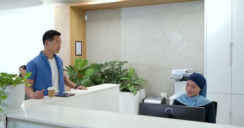Client holding coffee and tablet talking with receptionist in hijab at modern office desk