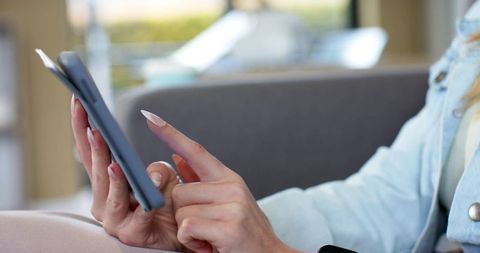 Woman tapping smartphone with long nails wearing denim jacket on grey sofa