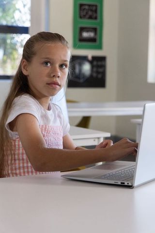 Preteen girl concentrating on laptop in bright classroom during remote learning session