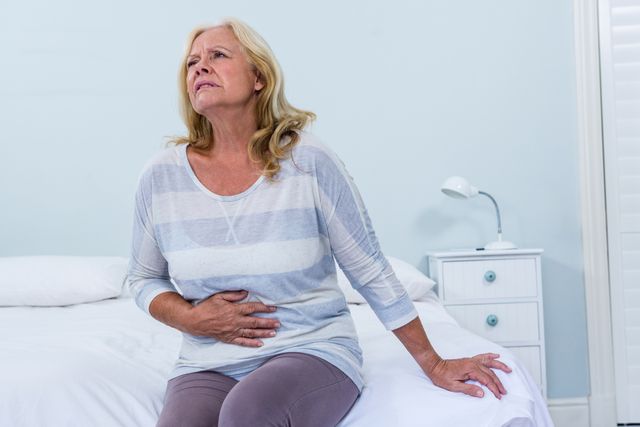 Senior woman sitting on her bed has stomach ache in bedroom