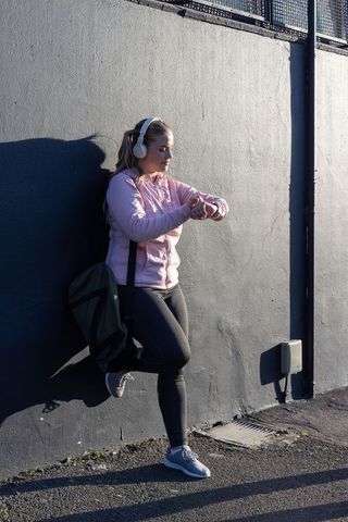 Teen woman leaning on urban wall checking smartwatch wearing headphones and backpack