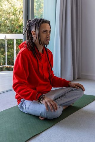 African american man meditating on yoga mat in red hoodie near open balcony door