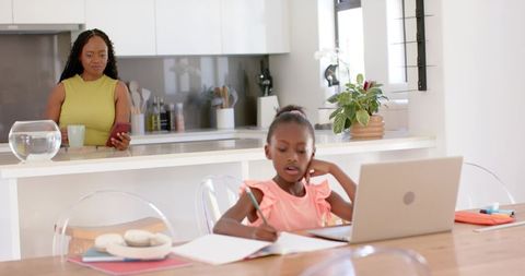 Mother monitoring daughter doing homework at kitchen table with laptop and notebooks