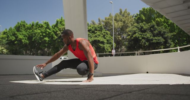 Front view of an athletic African American man in sportswear squatting, stretching his legs and holding his toes under a bridge while training on a sunny day in the city.
