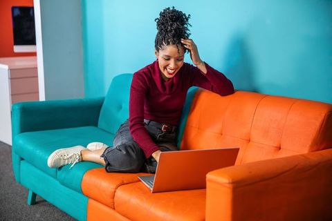 African american woman working on laptop on colorful teal and orange sofa, smiling