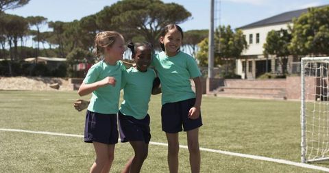 Three schoolgirls celebrating together on soccer field wearing mint tees and navy shorts