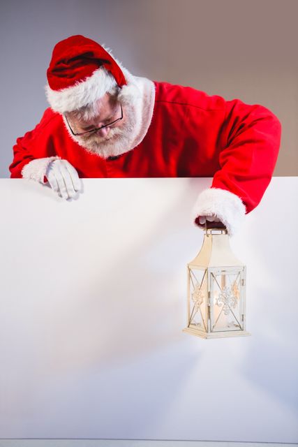 Santa Claus dressed in traditional red suit and hat holding a Christmas lantern while leaning on a white board. Ideal for holiday promotions, Christmas greeting cards, festive advertisements, and seasonal marketing materials.