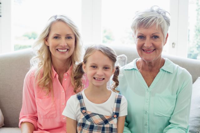 Three generations of women, including a grandmother, mother, and daughter, are sitting together on a sofa at home. They are smiling and appear happy, showcasing family bonding and togetherness. This image can be used for family-related content, advertisements, or articles focusing on multi-generational relationships and home life.