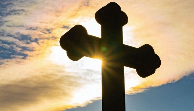 Stone trefoil cross silhouetting at sunset with sunburst halo and dramatic sky