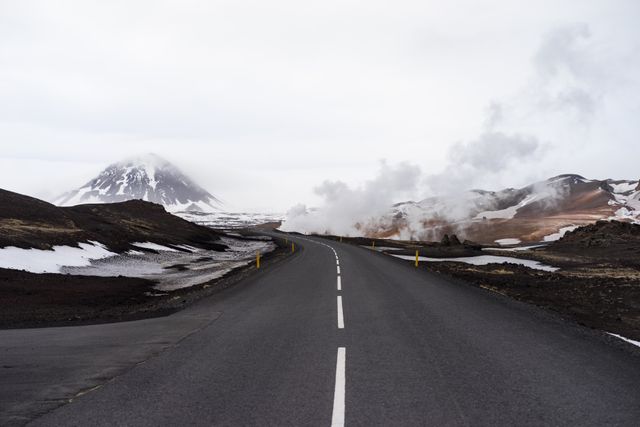 Image depicts an empty asphalt road leading through a barren volcanic landscape with steam rising from the ground and a snow-capped mountain in the distance. Sky is covered with clouds, contributing to the overall remote and rugged atmosphere. Perfect for travel brochures, adventure blogs, and nature documentaries focusing on adventurous journeys, geological formations, and remote landscapes.