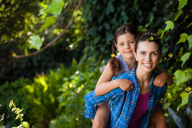 Portrait of cheerful woman piggybacking daughter against trees in backyard