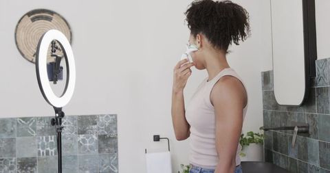 African american woman using facial skincare device in bathroom with ring light