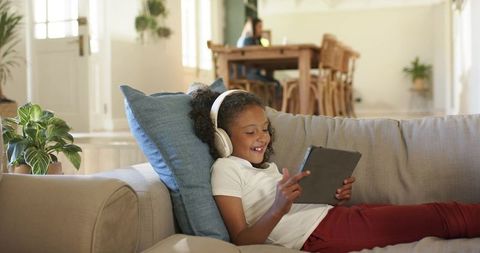 Smiling girl reclining on sofa with tablet and headphones enjoying learning at home