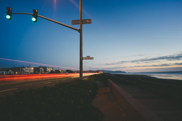 Scene captures a coastal highway during twilight with a traffic light overseeing the scenic road. Light trails of passing vehicles highlight the ongoing activity, blending with the serene ocean view on the right side of the frame. Recommended for use in travel magazines, promotional materials, or websites emphasizing coastal tourism, evening urban transportation, or scenic drives.
