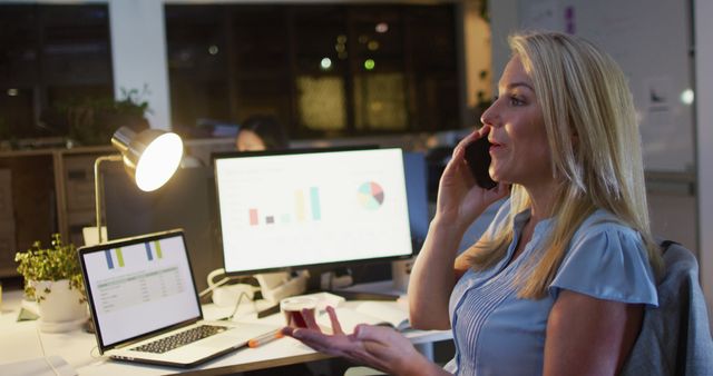 Professional businesswoman speaking on phone while working late in office. Her working environment includes multiple computer screens displaying charts and graphs, suggesting analysis or financial work. Ideal for use in articles about corporate life, business professionals, work-life balance, or modern office settings.