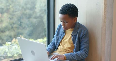 African american boy studying on laptop in cozy window seat with minimalist interior
