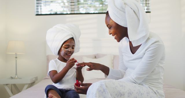 Happy african american mother and daughter sitting on bed and painting nails. staying at home in self isolation during quarantine lockdown.