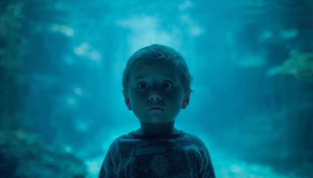 Child standing in teal-lit aquarium tunnel with wide-eyed cinematic gaze