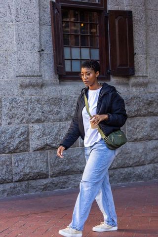 African woman walking with coffee past stone wall wearing puffer jacket and green crossbody bag