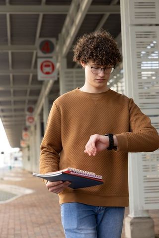 Teen checking smartwatch at bus stop holding notebooks commuting to school under overcast sky