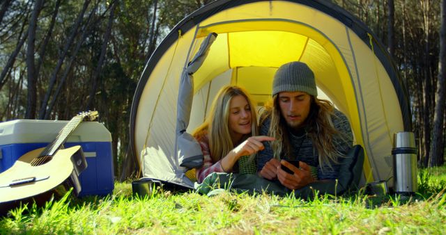 Young couple relaxing in a yellow camping tent, surrounded by green grass and trees. The scene includes a cooler and a guitar to the side, while the couple engages with a smartphone. Ideal for lifestyle, travel, outdoor adventure, camping equipment advertisements, or bonding activities themes.