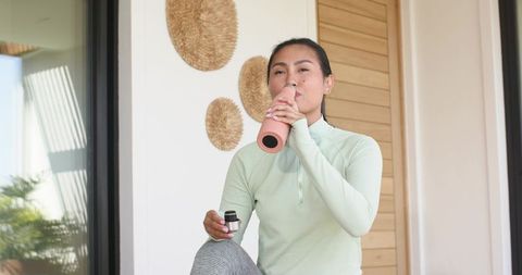 Asian woman sipping from matte pink reusable bottle while wearing mint activewear on porch