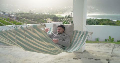 Indian man relaxing in striped hammock on coastal terrace using smartphone