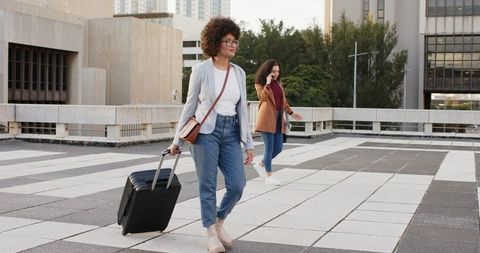 Two women walking across urban terrace pulling black wheeled suitcase, one talking phone