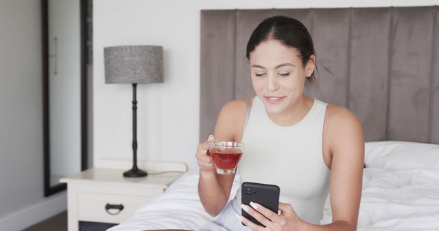 Young woman sitting on bed, drinking tea from clear glass cup while using smartphone, appearing relaxed and focused. A bedroom lamp on bedside table provides a cozy touch. Ideal for content related to modern lifestyle, morning routines, technology use, or leisure time at home.