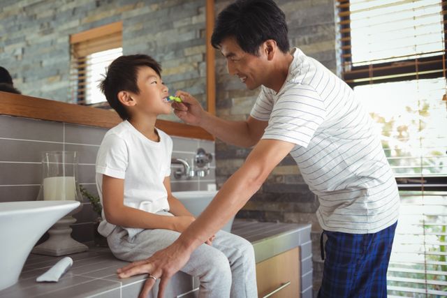 Asian father brushing his young son's teeth in a modern bathroom with stone walls. This stock photo is ideal for promoting family values, parenting tips, dental hygiene products, or home care routines. Perfect for websites, blogs, or advertisements focused on family life, health, and wellness.