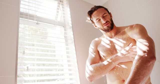 Young man is standing under the shower in a bright bathroom while sunlight filters through the window blinds. Droplets of water and bright light enhance focus on personal hygiene and self-care. Useful for websites or ads related to daily routines, wellness, health care, personal hygiene products, men's grooming, and home interior decor.