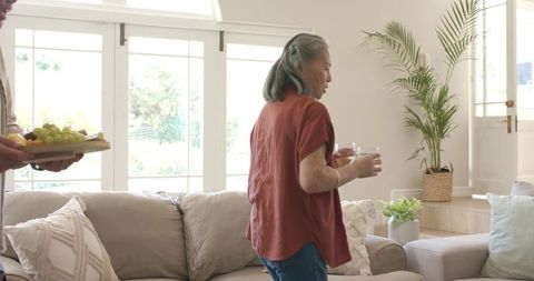 Mature woman serving drinks while partner offering charcuterie board in sunlit living room