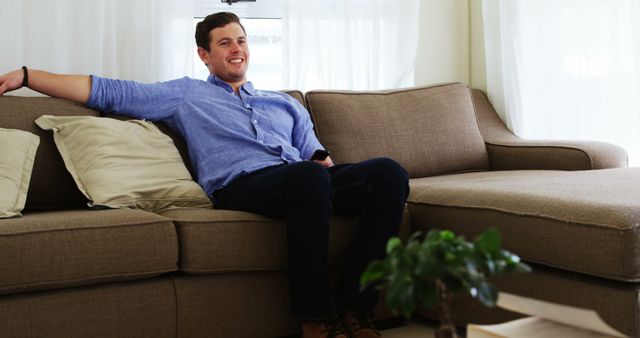 Young man sitting comfortably on a modern sofa in a bright living room with neutral decor. He appears relaxed and content, exuding a sense of ease and leisure. This image is perfect for promoting home decor, furniture, lifestyle blogs, and relaxation techniques.