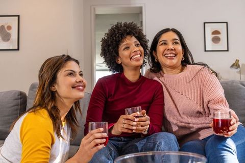 Multicultural friends laughing and relaxing on gray sofa while holding red drinks at home