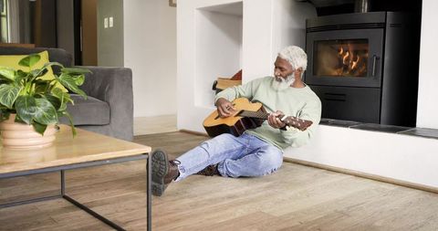Elderly man relaxing while playing guitar by fireplace