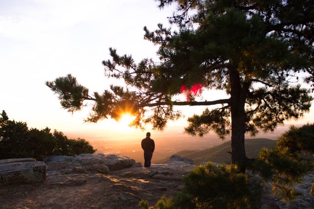 Silhouette of person standing on mountain overlooking a sunset. Useful for themes on solitude, adventure, and peaceful nature retreats. Perfect for websites, blogs, and social media posts about hiking, outdoor activities, and travel.