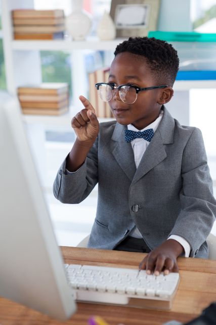 Young boy dressed in a suit and glasses, pointing while working at a desk with a computer. Ideal for concepts related to education, future leaders, young professionals, business training, and corporate environments.