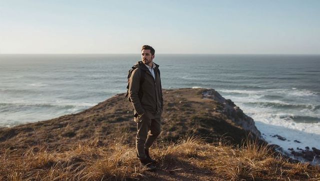 Solo hiker standing on coastal cliff overlooking ocean at golden hour wearing backpack