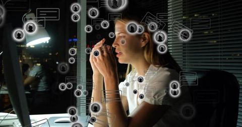 Working late woman leaning toward monitor holding glasses with digital chat overlays