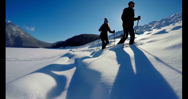 Caucasian couple enjoys a snowshoeing adventure in the wilderness. Their long shadows stretch across the pristine snow, highlighting a perfect winter day outdoors.