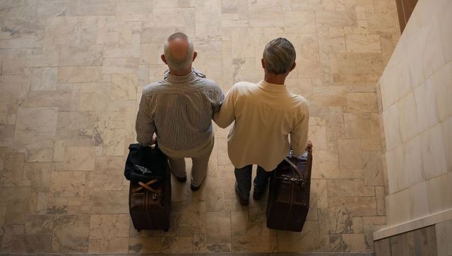 Two seniors standing side by side holding leather suitcases on marble foyer floor, travel companions