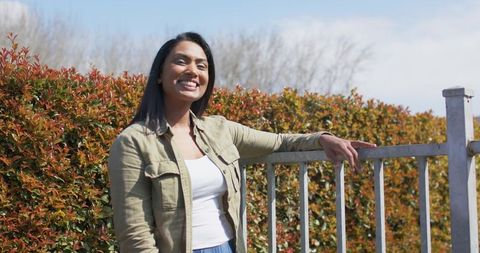 Indian woman smiling and leaning on railing in sunlit garden wearing khaki jacket